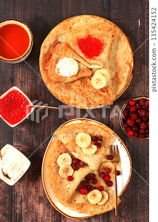 Delicious Russian pancakes with soft cheese, bananas, red caviar, and cranberries on the kitchen table. A healthy breakfast made with fresh ingredients, inspired by Maslenitsa Delicious Russian pancakes with soft cheese, bananas, red caviar, and cranberries on the kitchen table. A healthy breakfast made with fresh ingredients, inspired by Maslenitsa 135424152
