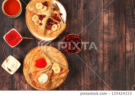 Delicious Russian pancakes with soft cheese, bananas, red caviar, and cranberries on the kitchen table. A healthy breakfast made with fresh ingredients, inspired by Maslenitsa Delicious Russian pancakes with soft cheese, bananas, red caviar, and cranberries on the kitchen table. A healthy breakfast made with fresh ingredients, inspired by Maslenitsa 135424153