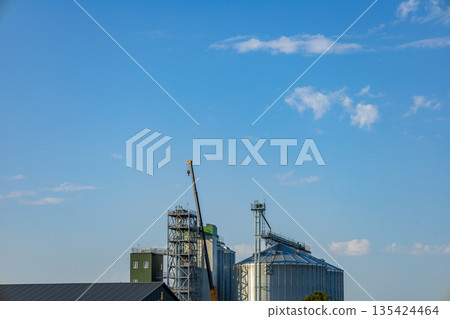 Various granary buildings and silos are seen with cranes working under a blue sky during the day Various granary buildings and silos are seen with cranes working under a blue sky during the day 135424464