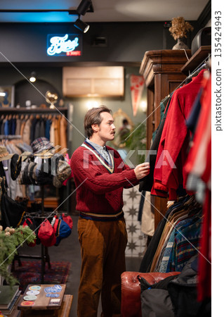 Stylish man customer picks trendy old fashioned outfit in fashionable selective second hand store. Stylish man customer picks trendy old fashioned outfit in fashionable selective second hand store. 135424943