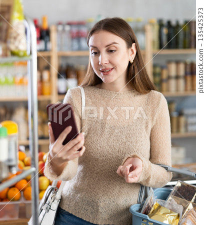 Happy young european girl talking to someone by video link during looking for something at store Happy young european girl talking to someone by video link during looking for something at store 135425373