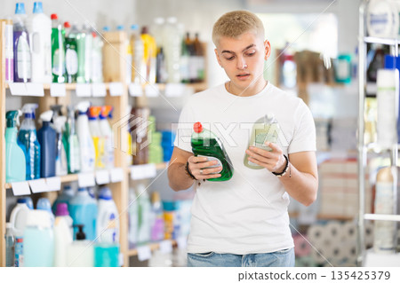 Attentive young man choosing between two dishwashing liquids standing between shelves in supermarket 135425379