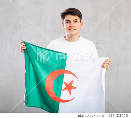 Reliant positive young man holds big national flag of Algeria with confident smile showing teeth. Reliant positive young man holds big national flag of Algeria with confident smile showing teeth. 135425420