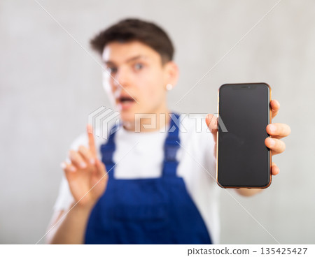 Unfocused man in jumpsuit shows close up empty mobile phone screen and points at device with finger Unfocused man in jumpsuit shows close up empty mobile phone screen and points at device with finger 135425427