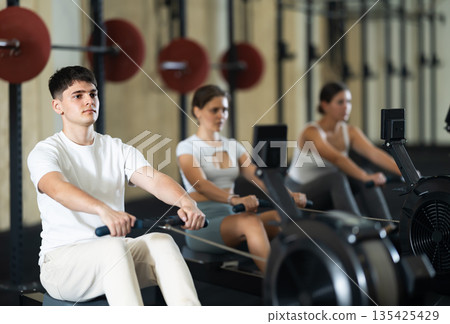 Young athletic guy in sportswear training on rowing machine in sport gym Young athletic guy in sportswear training on rowing machine in sport gym 135425429