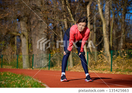 Woman standing on running track leaning forward with hands on knees after exercise 135425663