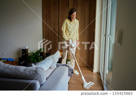 Woman standing in living room using cordless vacuum cleaner while dog sits on sofa 135425665