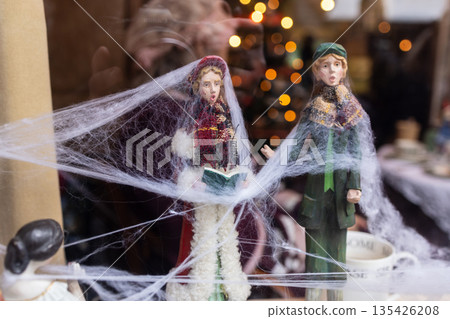 Two Victorian style caroler figurines covered in cobwebs stand near mugs and festive lights, evoking eerie nostalgia. Two Victorian style caroler figurines covered in cobwebs stand near mugs and festive lights, evoking eerie nostalgia. 135426208