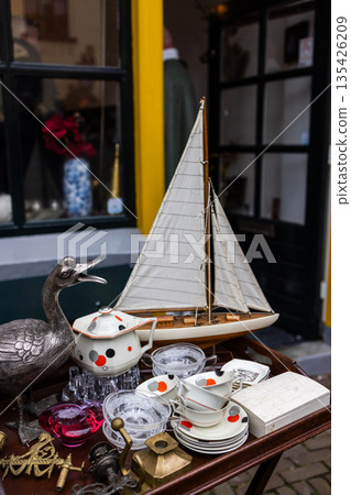 Outdoor table displays model sailboat with white sails, ceramic dishes with spiral motifs, glassware, and metal bird sculpture near shop entrance. 135426209