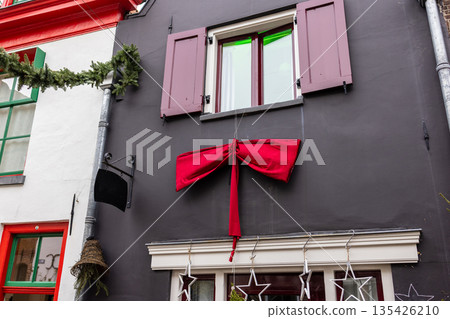 Dark gray facade with maroon shutters and red bow, festive stars and garlands adorn windows and roofline in holiday street scene. 135426210
