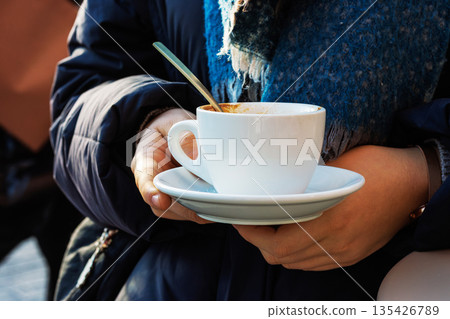 Young woman in winter coat holding empty coffee cup and saucer in street cafe 135426789