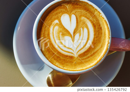 Cappuccino cup with heart latte art on a saucer with spoon, lit by natural sunlight Cappuccino cup with heart latte art on a saucer with spoon, lit by natural sunlight 135426792