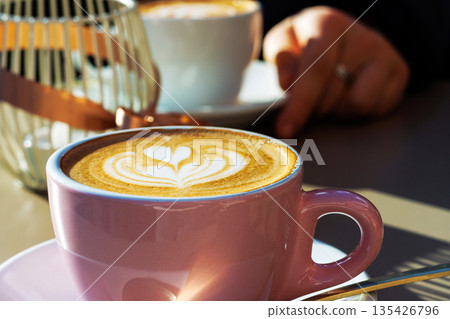 Two coffee cups with a heart latte art cappuccino on a cafe  table on a sunny day with a person sitting in the background. Selective focus 135426796