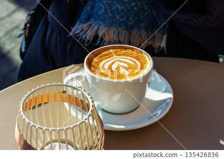 Sunlit cafe scene with a cup of cappuccino with latte art beside a candle holder decorated with ribbon. Hand of a person sitting in the background. Selective focus Sunlit cafe scene with a cup of cappuccino with latte art beside a candle holder decorated with ribbon. Hand of a person sitting in the background. Selective focus 135426836