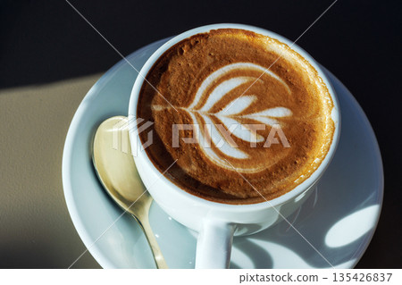 Cappuccino cup with heart latte art on a saucer with spoon, lit by natural sunlight 135426837