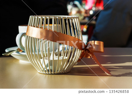 Sunlit cafe tabletop with a decorative candle holder wrapped with a ribbon bow, with a coffee cup and saucer softly blurred in the background. Sunlit cafe tabletop with a decorative candle holder wrapped with a ribbon bow, with a coffee cup and saucer softly blurred in the background. 135426858