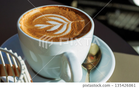 Sunlit close up of a cappuccino with latte art in a white cup on a saucer with a spoon on a cafe table. Sunlit close up of a cappuccino with latte art in a white cup on a saucer with a spoon on a cafe table. 135426861