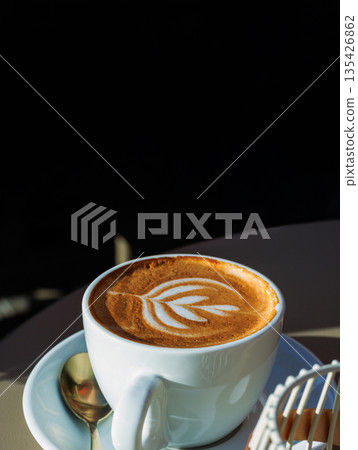 Vertical close up of a cappuccino with latte art in a white cup on a saucer with a spoon against a dark black background with large copy space. Vertical close up of a cappuccino with latte art in a white cup on a saucer with a spoon against a dark black background with large copy space. 135426862