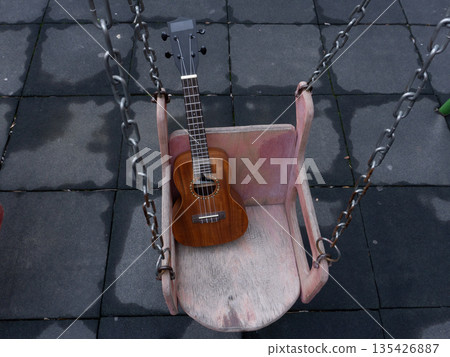 Acoustic ukulele resting on a wooden swing in a playground. Musical instrument outdoors on wet rubber tiles 135426887