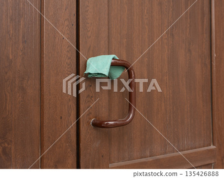 Green cleaning cloth draped over a brown wooden door handle. Housekeeping and hygiene concept. Domestic chore close up Green cleaning cloth draped over a brown wooden door handle. Housekeeping and hygiene concept. Domestic chore close up 135426888