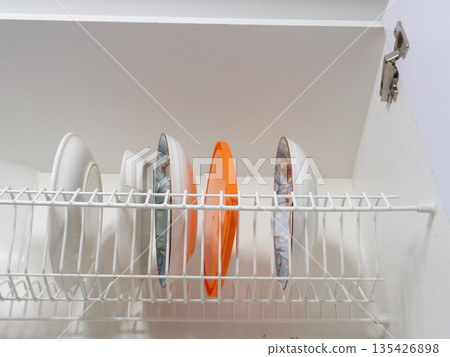 Dish drying rack inside a white kitchen cabinet. Wire shelf holding ceramic plates, bowls, and an orange plastic lid. Household organization and storage concept Dish drying rack inside a white kitchen cabinet. Wire shelf holding ceramic plates, bowls, and an orange plastic lid. Household organization and storage concept 135426898