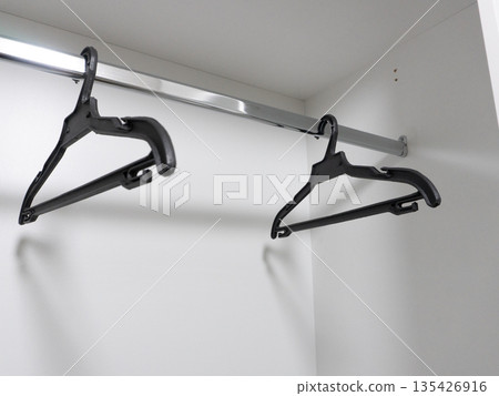 Two empty black plastic hangers on a chrome rod inside a white wardrobe. Clothing storage and organization background with copy space Two empty black plastic hangers on a chrome rod inside a white wardrobe. Clothing storage and organization background with copy space 135426916