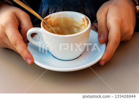 Hands holding an almost empty coffee cup on a saucer with a spoon, with visible latte residue in warm sunlight at a cafe table. Hands holding an almost empty coffee cup on a saucer with a spoon, with visible latte residue in warm sunlight at a cafe table. 135426944