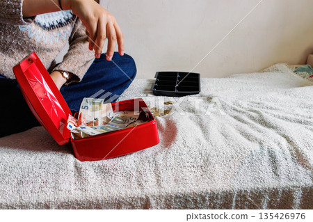 Indoor scene of a woman placing euro banknotes into an open red cash box on a bed covered with a textured blanket, with a coin tray nearby. Indoor scene of a woman placing euro banknotes into an open red cash box on a bed covered with a textured blanket, with a coin tray nearby. 135426976