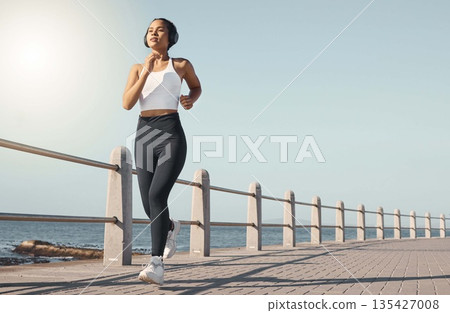 Fitness, woman run at beach and with headphones listening to radio with lens flare. Training or exercise, marathon or sportswear and female person running along the promenade listen to music 135427008