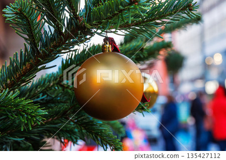 A gold christmas bauble hanging from a fir branch with a blurred street scene and people in the background. A gold christmas bauble hanging from a fir branch with a blurred street scene and people in the background. 135427112