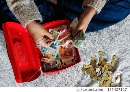 Top view of hands counting euro banknotes in open red cash box filled with cash, with a pile of euro coins on a textile bed blanket. Top view of hands counting euro banknotes in open red cash box filled with cash, with a pile of euro coins on a textile bed blanket. 135427117