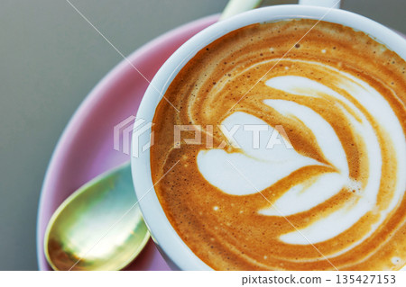 A closeup top view of a cappuccino topped with heart and flower shaped latte art in a white cup on a pink saucer with  a small spoon beside. Soft focus 135427153
