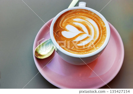 A cup of cappuccino with heart flower shaped latte art on a pink saucer with a small spoon. View from above on a smooth table surface. A cup of cappuccino with heart flower shaped latte art on a pink saucer with a small spoon. View from above on a smooth table surface. 135427154