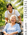 A foreign caregiver and an elderly Japanese patient stroll through the park in the soft sunlight, smiling. 135428741