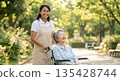 A foreign caregiver and an elderly Japanese patient stroll through the park in the soft sunlight, smiling. 135428744