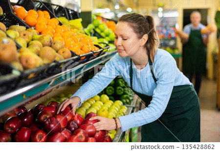 Friendly middle-aged female seller in apron arranging red apples in supermarket 135428862