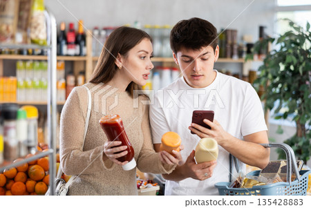 Positive young woman and man looking to buy sauce, scans QR code from label in food supermarket 135428883