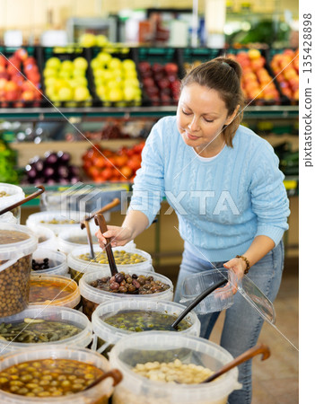 Middle-aged European female picking up olives with scoop from plastic container in grocery store Middle-aged European female picking up olives with scoop from plastic container in grocery store 135428898