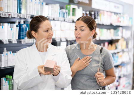 Female pharmacist advises a woman visitor a good skin care remedy, standing in the sales hall of pharmacy Female pharmacist advises a woman visitor a good skin care remedy, standing in the sales hall of pharmacy 135428938