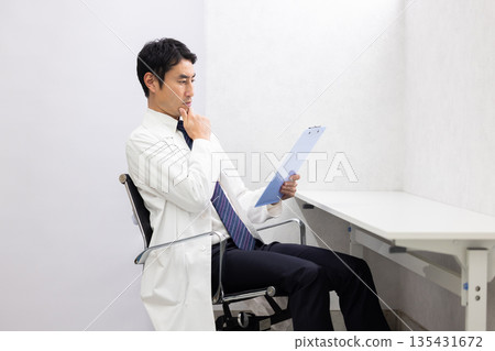 一位身穿白大褂的男醫生神情嚴肅地閱讀著一份病歷。 一位身穿白大褂的男醫生神情嚴肅地閱讀著一份病歷。 135431672