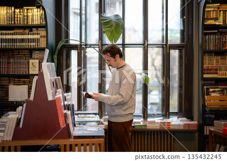 Mindful man choosing bestselling books in bookstore surrounded by shelves filled with publications. Mindful man choosing bestselling books in bookstore surrounded by shelves filled with publications. 135432245
