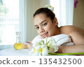 Beautiful woman relaxing on spa bed with plumeria flowers and massage oil, smiling gently in peaceful atmosphere, representing wellness, aromatherapy, skincare, and luxury spa treatment experience 135432500