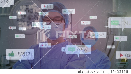 Male surgeon and scrub nurse reviewing monitors under surgical light in OR, with social icons Male surgeon and scrub nurse reviewing monitors under surgical light in OR, with social icons 135433166