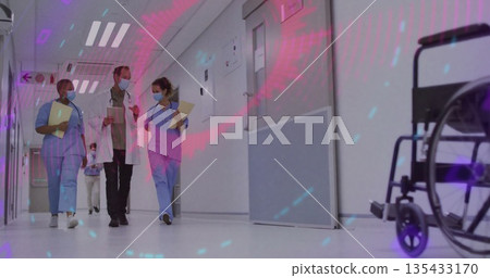 Walking team in scrubs and lab coat down hospital corridor, with tablet, stethoscope, clipboards 135433170