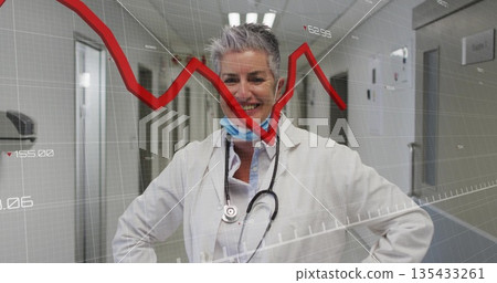 Smiling senior female doctor wearing lab coat and stethoscope in hospital corridor, with red graph 135433261