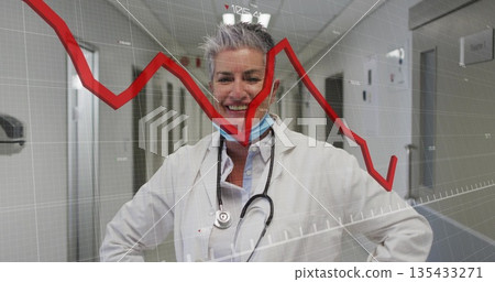 Standing doctor wearing coat and blue mask with stethoscope in hospital corridor, showing red graph Standing doctor wearing coat and blue mask with stethoscope in hospital corridor, showing red graph 135433271