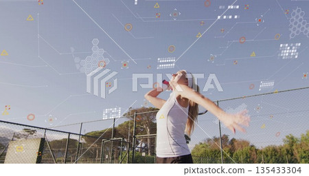 Serving female tennis player in tennis attire tossing ball swinging racket at net on outdoor court 135433304