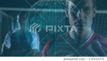 Red-shirted player holding soccer ball on pitch, with net and wireframe globe overlay, copy space Red-shirted player holding soccer ball on pitch, with net and wireframe globe overlay, copy space 135433371