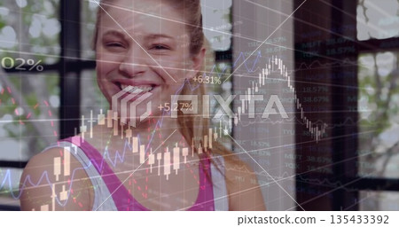 Smiling woman wearing pink-white tank top analyzing candlestick charts in studio with grid windows 135433392