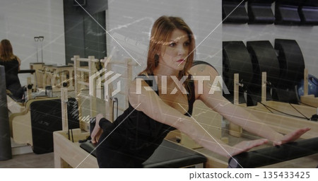 Performing Pilates exercise woman wearing black sportswear in Pilates studio, with reformer, mirror Performing Pilates exercise woman wearing black sportswear in Pilates studio, with reformer, mirror 135433425